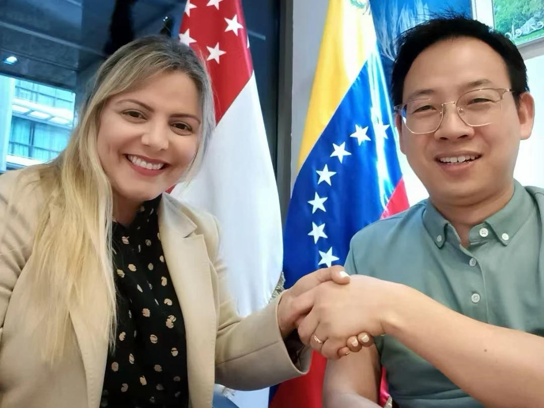 Participants exchange a handshake with Venezuelan and Singaporean flags behind them.