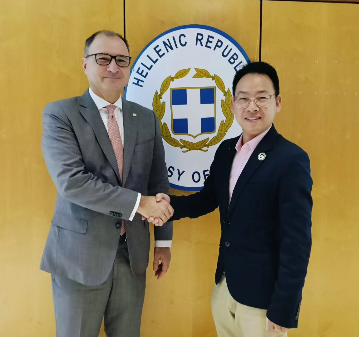 Participants exchange a handshake in front of the Hellenic Republic embassy emblem.