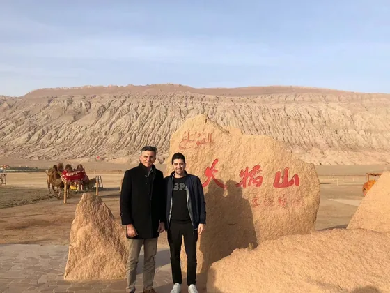Two participants beside a landscape marker in a dry, mountainous setting.