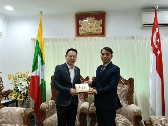 Participants exchange a presentation item in front of Myanmar and Singapore flags.